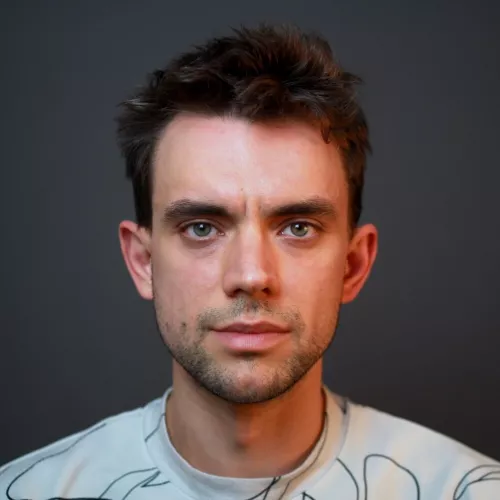 Portraitfoto eines weißen Manns mit kurzen braunen Haaren und leichten Barstoppeln. Er trägt ein weißes T-Shirt vor einem grauen Hintergrund.