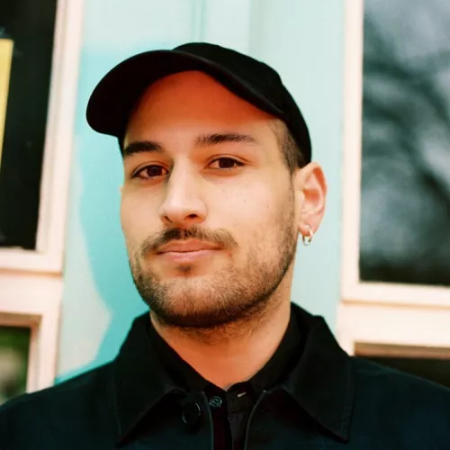Es ist der Kopf von Person mit schwarzer Kappe und Hals und Ansatz von einem schwarzen Pulli vor einem verschwommenem Hintergrund zu sehen.