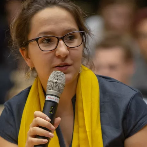 Eine Frau mit Brille, hellbraunen Haaren und einem gelben Schal, sie hat ein Mikrophon in der Hand und schaut jemanden neben ihr an, hinter ihr ist ein Publikum zu erkennen, es ist verwischt.