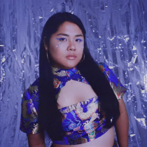 Das Foto ist ein Portrait von Vietnamesisch-Deutscher Sängerin und Produzentin veenus. Die Gesamtstimmung ist verträumt durch einen schimmernden, glitzernden Hintergrund in mattblau. In der Mitte des Bildes schaut veenus ernst in die Kamera. Sie hat lange schwarze Haare und ein vietnamesisch traditionell inspiriertes Oberteil aus Seide an. Die Seide ist mit goldenen Drachen bestickt, das typisch vietnamesische Design zeigt einen Halskragen. 
