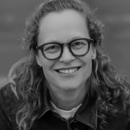 black and white portrait photo of a woman, smiling, she wears glasses and has long curly hair