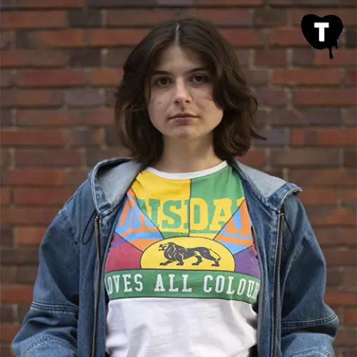 Özge, eine junge Frau mit rundem Gesicht und kurzen braunen Haaren, steht in einem bunten Shirt und Jeansjacke vor einer Ziegelsteinmauer.