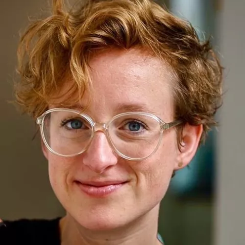 Das Bild zeigt das Gesicht einer Frau mit kurzen blonden Haaren und einer Brille, die lächelnd in die Kamera blickt.