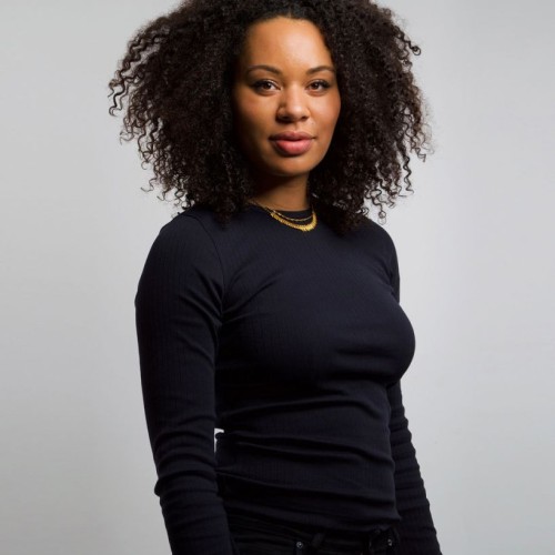 A portrait image of a German-Jamaican brown woman with an afro-like hairstyle wearing a black long sleeve top.